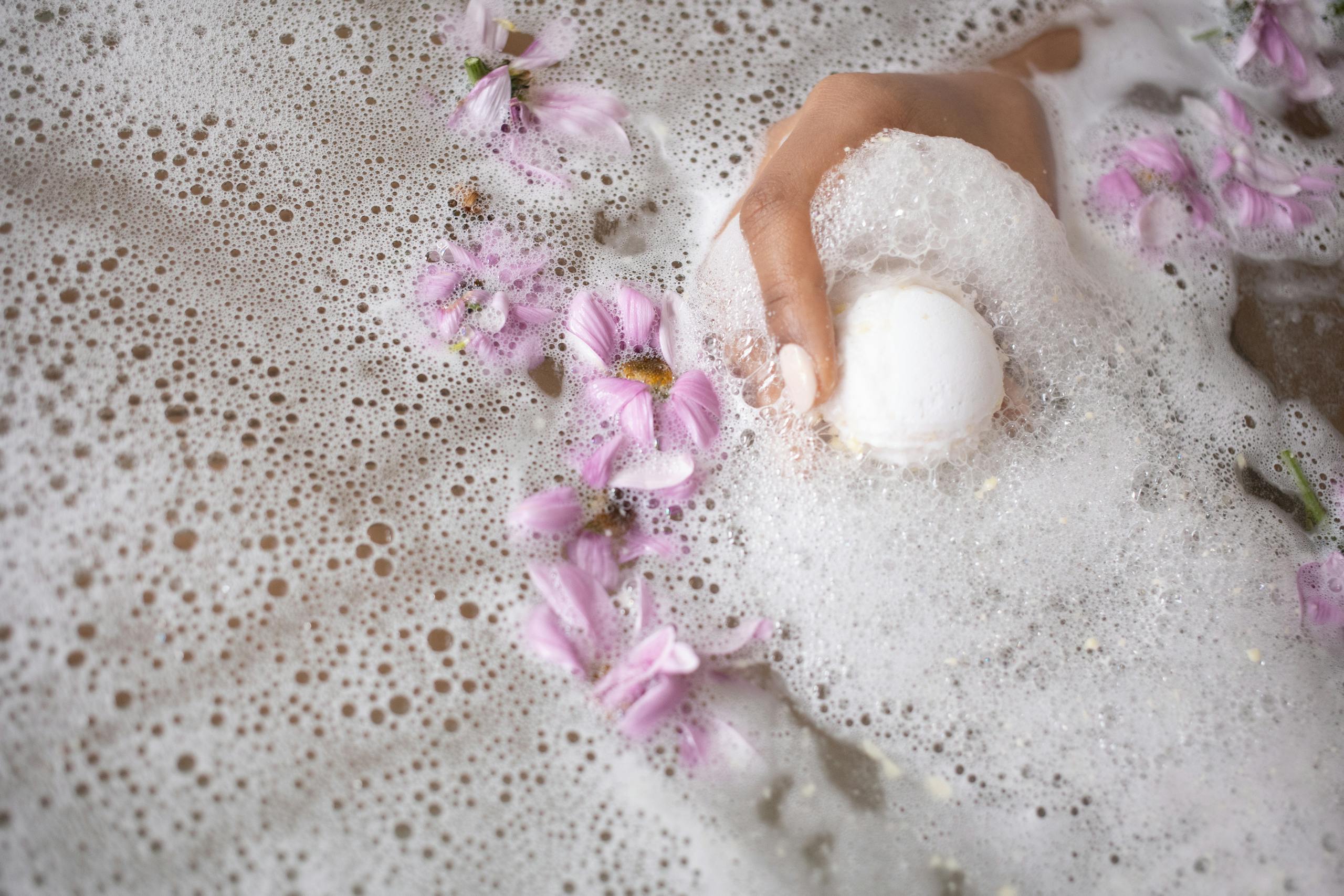 A calming spa scene with a hand holding a bath bomb amidst pink flower petals.